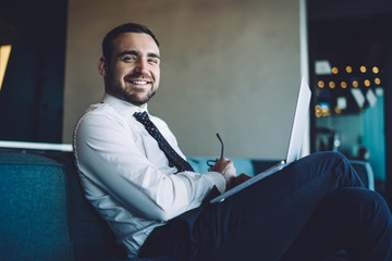 Portrait of cheerful male expert sitting at couch with laptop device and smiling at camera while...