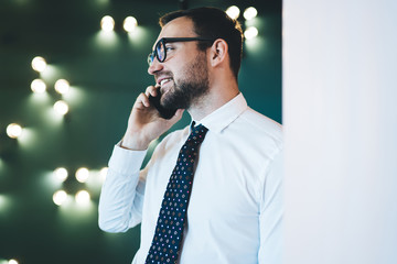 Side view of successful cheerful man director enjoying conversation using 4g connection on digital smartphone, male economist talking via cellular while standing in corridor company during break at jo