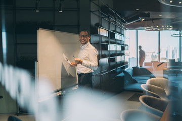Half length portrait of cheerful male corporate worker in optical spectacles holding report in hand and smiling at camera near flipchart, cheerful businessman planning startup project in workspace