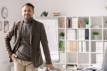 Confident mature architect in formalwear standing by desk in office