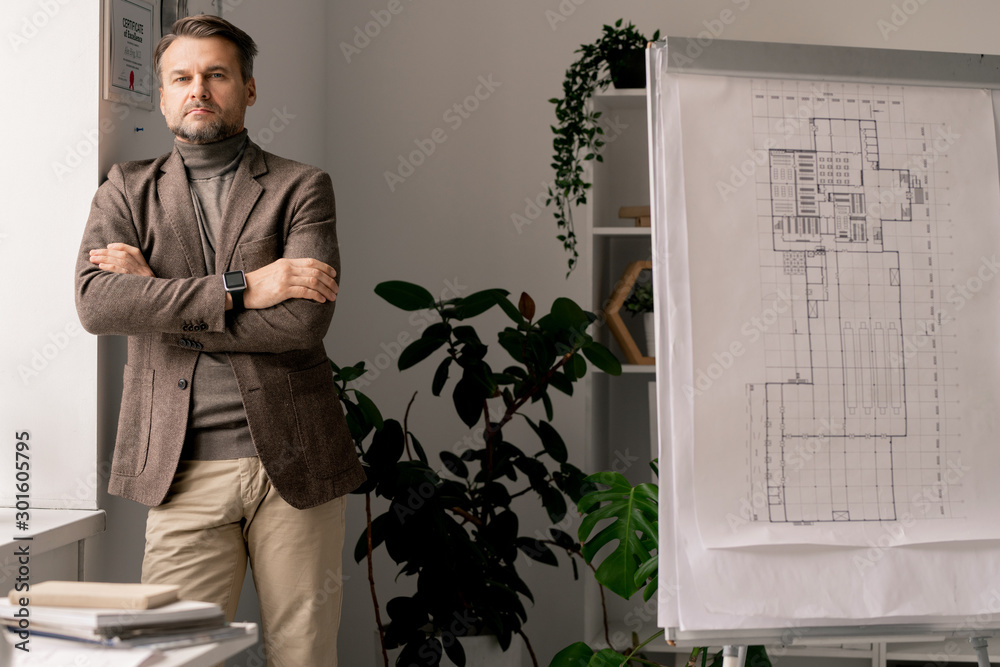 Poster serious architect or engineer with his arms crossed on chest standing by window