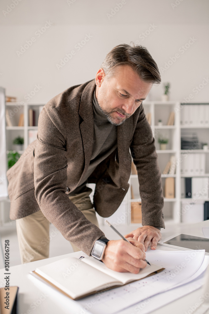 Poster Mature architect bending over open notebook on desk while making working notes