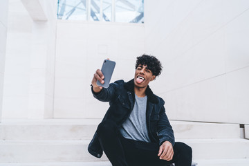 Joyful curly black guy taking selfie and showing tongue