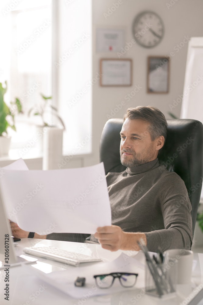 Poster Mature confident architect looking at sketches on papers while sitting by desk