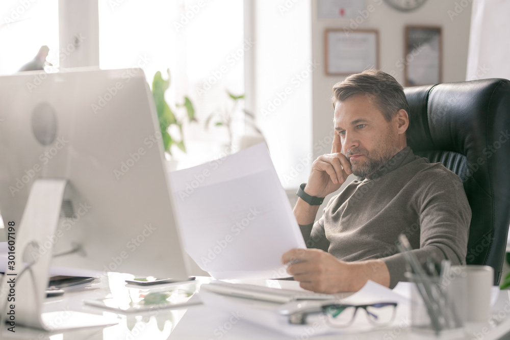 Poster mature serious architect looking at sketches while checking them by workplace