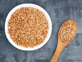 Buckwheat groats in a ceramic white bowl on a dark concrete background with wooden spoon. Healthy eating concept