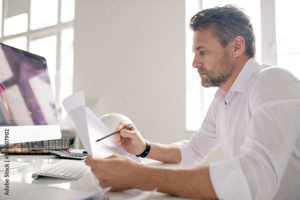 Poster mature serious engineer in white shirt pointing at sketch while checking it
