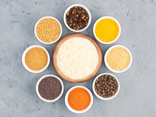 Allspice, pepper chili, fenugreek, cloves, turmeric, masala, dry ginger, mustard seeds around rice bowl on gray concrete background. Indian cuisine, ayurveda, naturopathy concept