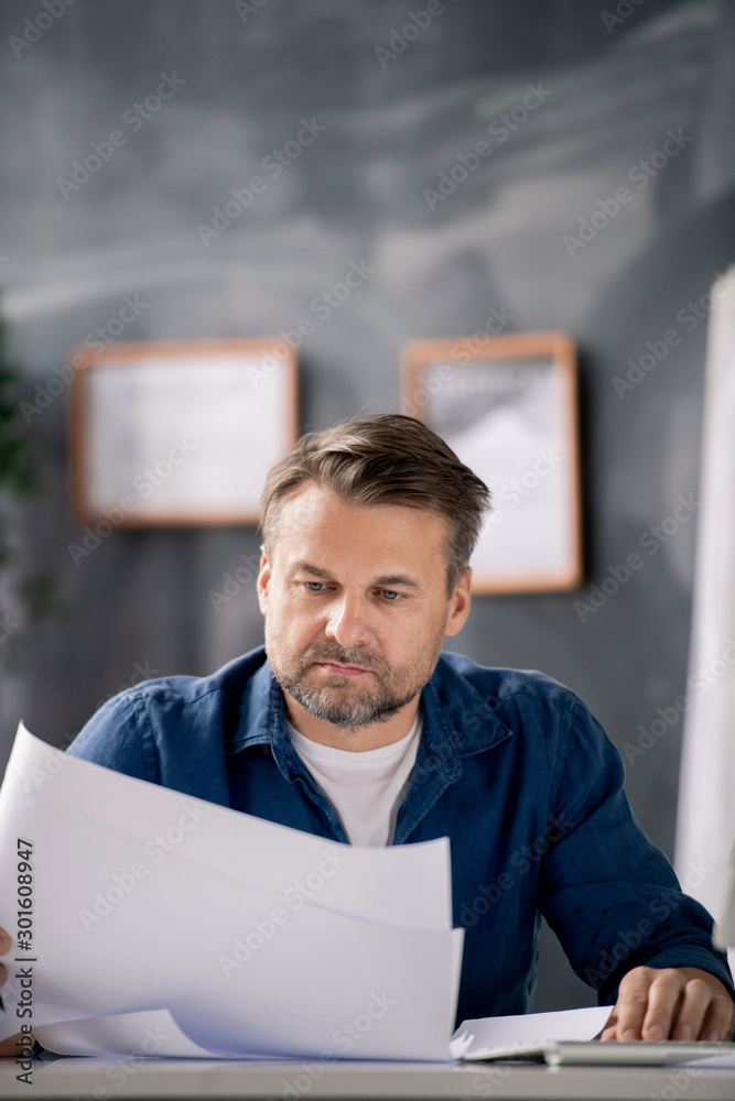 Poster bearded middle aged architect in casualwear looking at blueprint with sketch