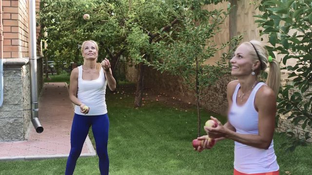 Two blonde Twins in white t-shirts juggle apples in the fresh air on the green grass.