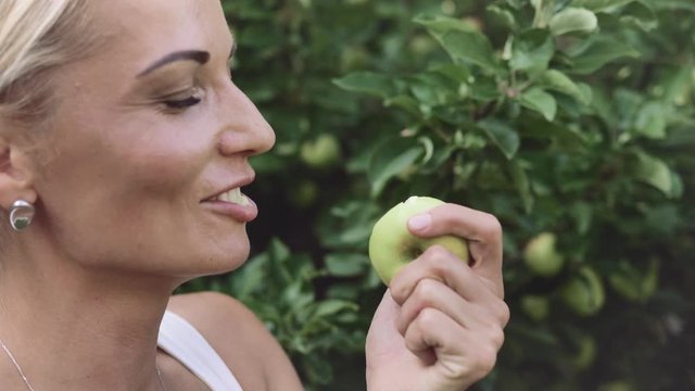 Beautiful young blonde girl bites off a green apple and winks at the camera, close-up