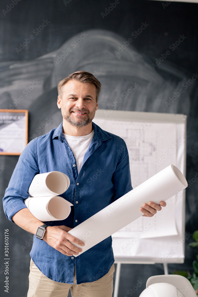 Poster successful mature engineer with rolled blueprints standing in front of camera