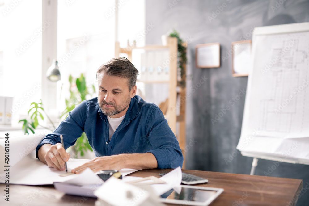 Poster mature architect in casualwear drawing line on blueprint while making sketch