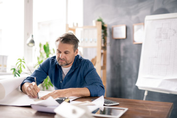 Mature architect in casualwear drawing line on blueprint while making sketch