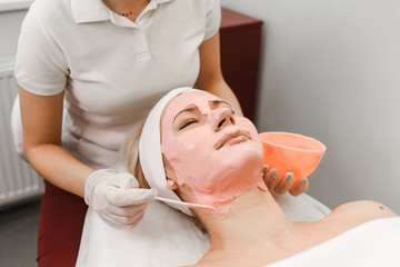 Obraz premium Doctor beautician applies anti-aging mask to woman face. Girls lies on a cosmetology procedure. Facial mask close-up