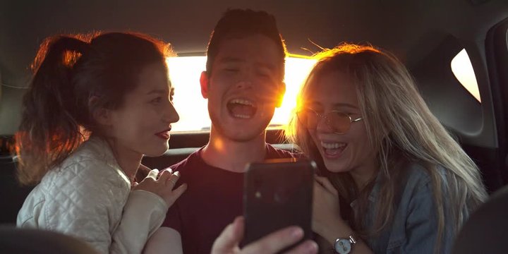 Fun Young Womens and man laugh and do a selfie In Back Seat Of Moving Car. Slow Motion. Sunset on background. flares