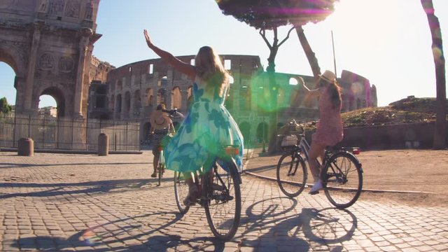 Three happy young women friends tourists riding bikes at Colosseum in Rome, Italy at sunrise.