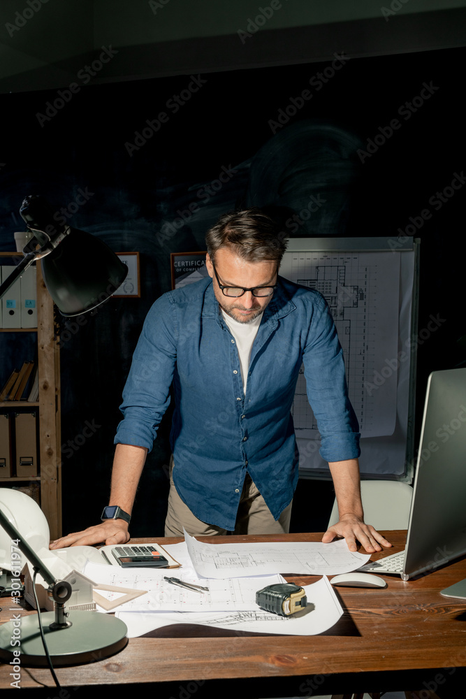 Poster mature engineer looking at sketch on paper during overtime work at night