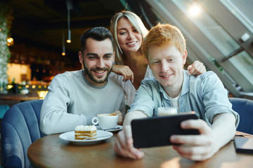 Fototapeta na wymiar Two happy guys and pretty blonde girl making selfie on smartphone in cafe