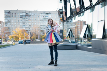 Happy young mother with little daughter on the arms and shopping bags in hand. Shopping day. Mall on background