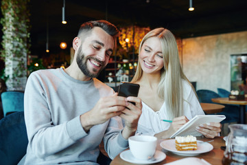 Young cheerful couple looking at smartphone screen while sitting in cafe