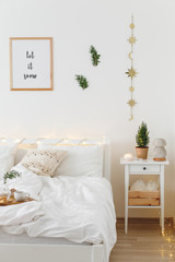 Christmas or new year winter home interior decor. Holiday decorated room. White stylish cozy scandinavian bedroom with little christmas tree, coniferous twigs or pine branches and led garland lights.