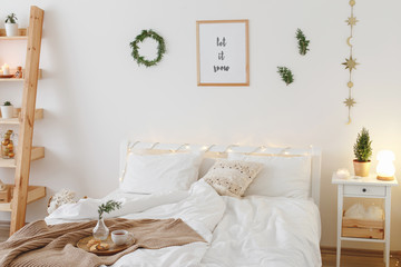 New year winter home interior decor. Christmas holiday decorations: little christmas tree, wreath, coniferous twigs, pine branches, led garland lights, blanket. White stylish cozy scandinavian bedroom