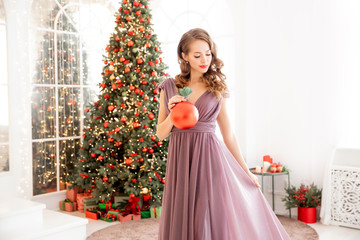 Christmas tree decoration, beautiful girl holding red ball in her hands, light from window. Concept party in evening dress