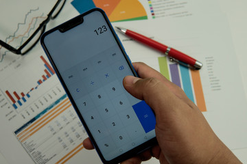 Fototapeta premium Adult male using phone as calculator with graph chart report document, pen and eye glasses on background. Finance banking concept.