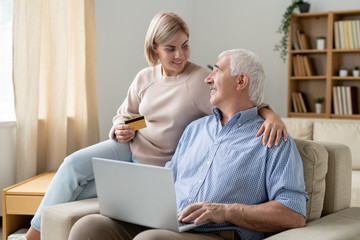 Pretty casual woman with credit card looking at her retired father with laptop