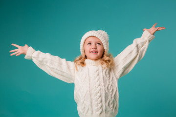 little blonde girl in white winter knitted hat and sweater on blue background isolate, space for...