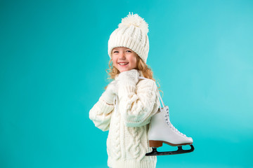 little blonde girl smiling in a winter white hat and sweater, holding skates. isolate on blue...