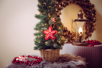 Small portable tabletop decorated Christmas tree on table, party lights illuminated, red felt fabric ornament. Light bulb lantern with pine cone wreath on background.