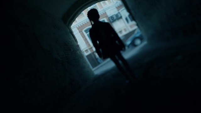 blurred young girl silhouette with Finnish braids walks forward camera in dark gloomy street gateway tilt shot