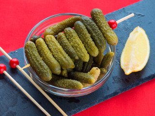 Gherkins in a crystal bowl on a slate table with toothpicks and lemon in a red background.