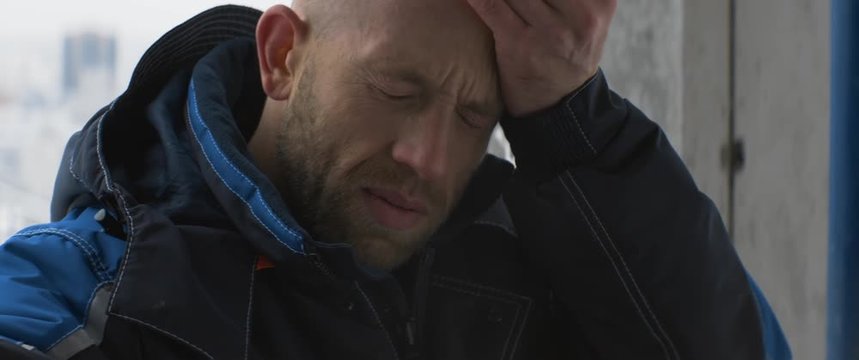 Tired caucasian worker or foreman in blue uniform takes off white helmet and wipes sweat from his forehead inside an unfinished building. Fatigue, feeling unwell, sadness, anamorphic shot