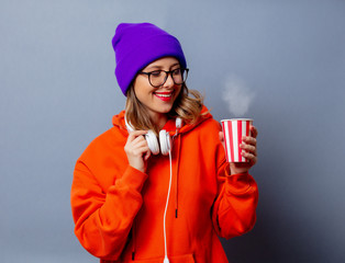 Style girl in orange hoodie and purple hat with cup on grey background