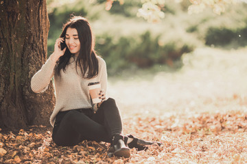 Beautiful woman using phone and drink coffee sitting near tree in autumn park.