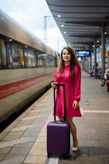 Young woman dressed in red dress staying with big purple case at railway station, waiting for her train to come to the platform