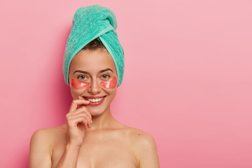 Glad European woman takes care of delicate skin around eyes, applies collagen patches, wears minimal makeup, wrapped bath towel on head, stands naked against pink background. Beauty concept.