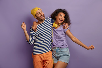 Fototapeta premium Friendly mixed race woman and man embrace and dance joyfully, spread hands, enjoy lovely music, feel energetic, dressed in fashionable clothes, isolated over bright purple background. Happy emotions