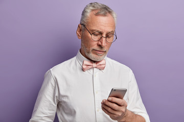 Elegant senior man works on his phone, concentrated in display tries to understand how to use modern technologies, wears round optical glasses, white formal shirt with bowtie, isolated on purple wall