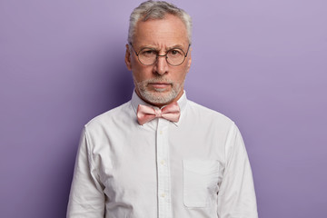 Portrait of handsome grey haired European man looks with strict face expression through round glasses, wears formal white shirt and bowtie, analyzes information, has wise look, stands indoor