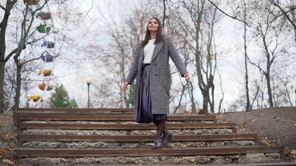 A young girl dancing in a coat on the street in autumn. The camera watches a girl in a warm coat walking and dancing on a sunny autumn street, she smiles and laughs and rejoices.