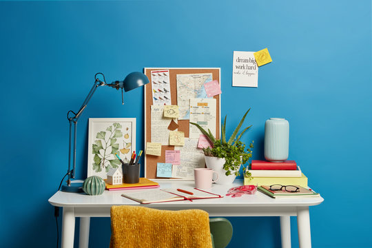 Work place of student. Desktop with lamp, opened notebook, stationery and green indoor plant, mug of coffee sticky notes on blue wall with written motivation phrases and special reminders. Home office