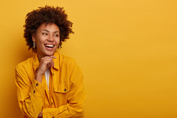 Fototapeta na wymiar Photo of cheerful African American woman with natural beauty, carefree expression, looks aside, has happy friendly attitude, talks casually with friend, wears bright jacket, isolated over yellow wall