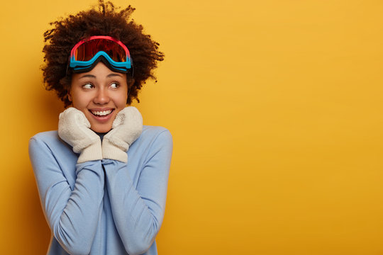 Ski resort and snowboarding. Glad smiling dark skinned woman wears white gloves, wears ski goggles and blue turtleneck, looks aside, stands over yellow background, dreams about nice family vacation