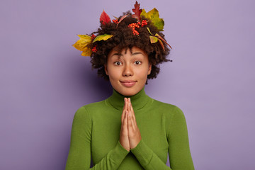 Pretty young Afro American woman asks for favour, keeps palms together, prays sincere, has autumn leaves in hair, wears green jumper, asks for help, says namaste, isolated over purple background