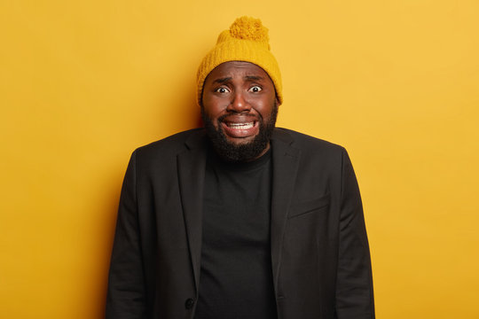 Nervous Awkward Black Man With Thick Beard, Feels Bothered, Admits His Mistake, Clenches Teeth, Wears Yellow Knitted Hat And Formal Suit, Scared Of Bad Consequences, Hears Terrible News, Isolated