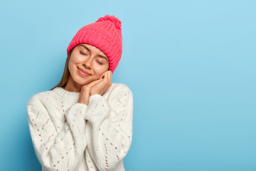 Photo of gentle young woman closes eyes, tilts head, imagines something pleasant, wears pink hat, winter sweater, poses against blue background, copy space. Pleasant emotions and feelings concept